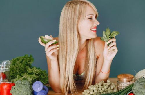 A smiling woman holding avocado and basil, surrounded by fresh vegetables and fruits for healthy eating.